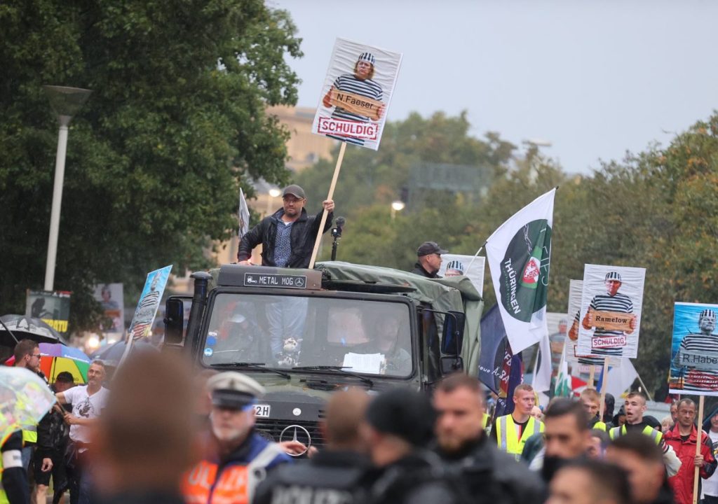 Tag der Deutschen Einheit in Gera 2023: Versammlungsanmelder Christian Klar steht auf einem historischen Militärfahrzeug und hält ein Plakat von Bundesinnenministerin Faeser in Sträflingskleidung und der Überschrift „schuldig“ in der Hand bei einer Demonstration des Bündnisses „Miteinanderstadt Gera“.